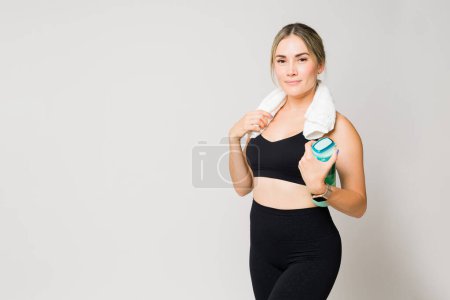 Téléchargez les photos : Femme athlétique finissant son entraînement à l'aide d'une serviette de gym et d'une bouteille d'eau devant un arrière-plan de studio avec espace de copie - en image libre de droit