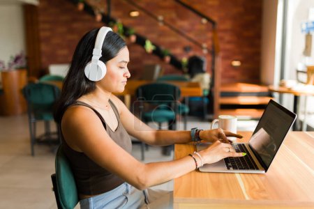 Téléchargez les photos : Jeune étudiante hispanique avec écouteurs écoutant de la musique et faisant des devoirs et travaillant au café - en image libre de droit