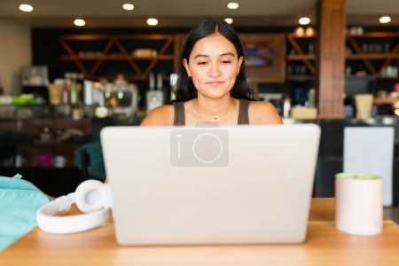 Téléchargez les photos : Belle jeune femme hispanique faisant du travail à distance en utilisant son ordinateur portable tout en prenant un café dans un café - en image libre de droit
