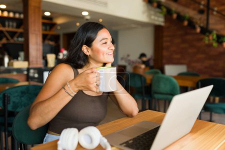 Téléchargez les photos : Hispanique heureuse étudiante au café boire du café et sourire tout en faisant du travail à distance en utilisant son ordinateur portable - en image libre de droit