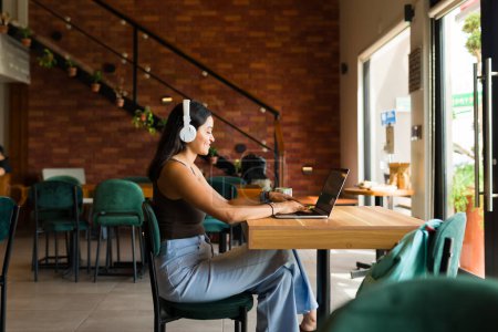 Téléchargez les photos : Profil d'une femme latino heureuse souriante avec écouteurs utilisant la zone wifi au café et travaillant sur l'ordinateur portable - en image libre de droit