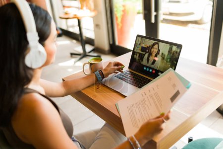 Téléchargez les photos : Vue arrière d'une jeune femme avec un casque dans un café ayant un appel vidéo pour un entretien d'embauche tout en utilisant son CV - en image libre de droit