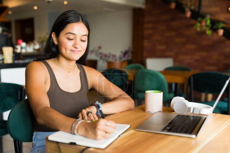 Téléchargez les photos : Belle femme hispanique écrit dans le carnet ou l'ordre du jour tout en étudiant et en faisant des devoirs à l'aide de l'ordinateur portable dans le café - en image libre de droit