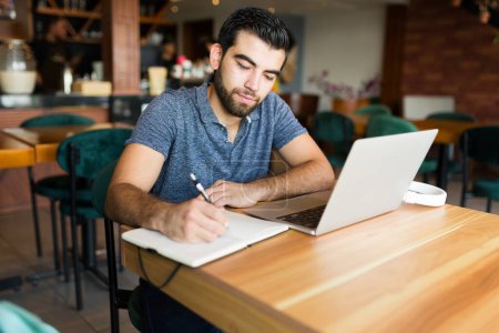 Téléchargez les photos : Beau latin homme et entrepreneur écrit à l'ordre du jour tout en travaillant à distance sur l'ordinateur portable au café - en image libre de droit