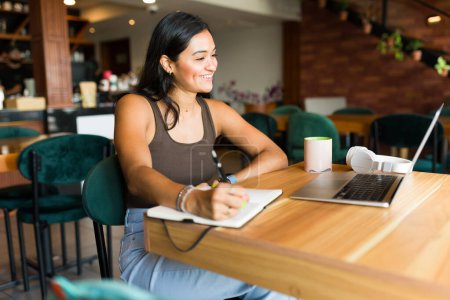 Téléchargez les photos : Femme hispanique excitée et entrepreneure riant tout en prenant des notes sur son agenda lors d'un appel vidéo de travail sur l'ordinateur portable - en image libre de droit