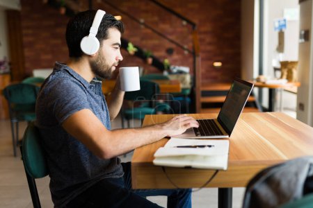 Téléchargez les photos : Vue latérale d'un homme latin avec écouteurs buvant du café au café et faisant du travail à distance à l'aide de son ordinateur portable et de la zone Internet - en image libre de droit