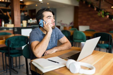Téléchargez les photos : Homme hispanique occupé entrepreneur dans le café parler lors d'un appel de travail au téléphone et faire du travail à distance sur l'ordinateur portable - en image libre de droit