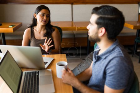 Téléchargez les photos : Jeune femme hispanique parlant avec un homme indépendant et travaillant ensemble en utilisant les ordinateurs portables au café - en image libre de droit