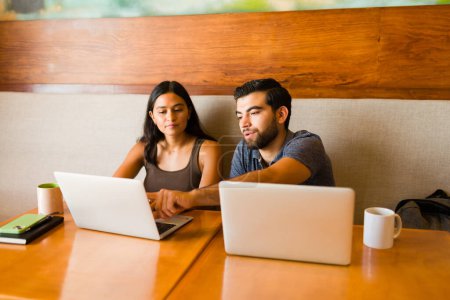 Téléchargez les photos : Homme hispanique intelligent aidant une jeune femme travaillant sur l'ordinateur portable tout en faisant du travail indépendant ensemble au café - en image libre de droit