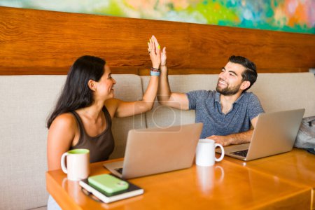 Téléchargez les photos : Femme et homme joyeux faisant un high five et célébrant un travail d'affaires réussi tout en travaillant à distance au café - en image libre de droit