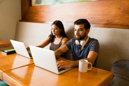 Téléchargez les photos : Couple hispanique en utilisant leurs ordinateurs portables et la zone Internet du café tout en faisant du travail indépendant à distance - en image libre de droit