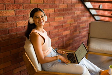 Téléchargez les photos : Joyeux latino femme avec casque souriant en utilisant son ordinateur portable et la zone wifi dans un beau café - en image libre de droit