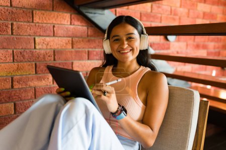 Téléchargez les photos : Femme heureuse souriant en utilisant sa tablette et son crayon dans la zone wifi du café tout en écoutant de la musique avec des écouteurs - en image libre de droit