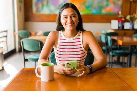 Téléchargez les photos : Belle femme latine souriante regardant la caméra tout en envoyant des SMS sur le smartphone au café et en buvant du café - en image libre de droit