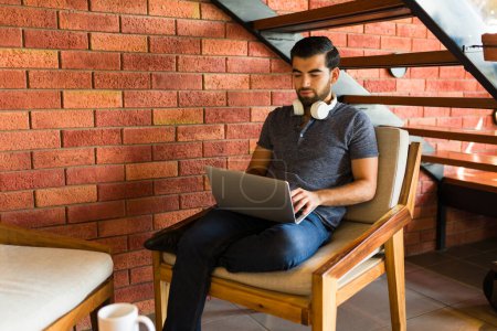 Téléchargez les photos : Mexicain intelligent homme et pigiste avec écouteurs faire du travail à distance en utilisant son ordinateur portable relaxant dans un café - en image libre de droit