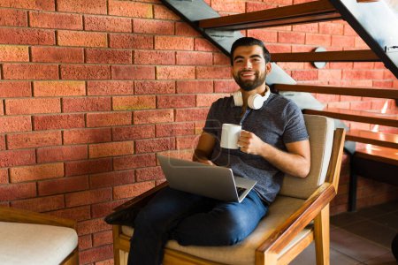Foto de Feliz hombre latino sonriendo bebiendo una taza de café mientras trabaja usando su portátil en la zona de internet de la cafetería - Imagen libre de derechos