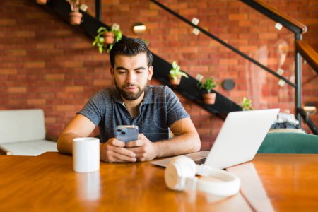 Photo for Handsome young man texting on the smartphone while doing remote freelance work on the laptop and drinking coffee - Royalty Free Image