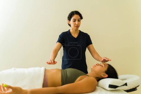 Photo for Hispanic therapist giving a reiki treatment to a relaxed young woman at the wellness center getting an alternative energy body massage - Royalty Free Image
