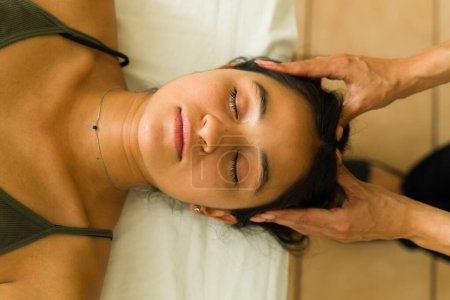 Téléchargez les photos : Vue du dessus de la femme latine détendue avec les yeux fermés au spa relaxant tout en obtenant un reiki alternatif ou massage de la tête - en image libre de droit