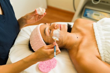 Téléchargez les photos : Femme hispanique détendue recevant un masque facial et un traitement d'exfoliation au spa de beauté tout en se relaxant et en se reposant - en image libre de droit
