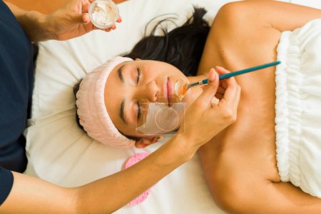 Foto de Hermosa mujer latina recibiendo una máscara facial y tratamiento de cuidado de la piel mientras hace exfoliación en el spa - Imagen libre de derechos