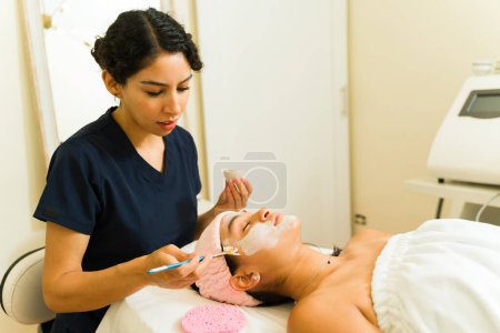 Téléchargez les photos : Jeune femme travaillant au spa de beauté donnant un soin de la peau soin du visage à une jeune femme relaxante - en image libre de droit