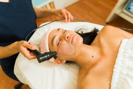 Foto de Atractiva mujer hispana en el spa de belleza recibiendo un tratamiento facial con láser para prevenir el envejecimiento de la piel y lucir relajada - Imagen libre de derechos