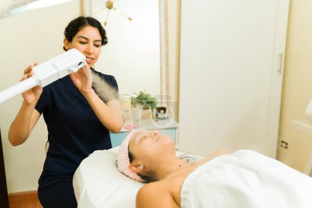 Téléchargez les photos : Femme heureuse donnant un traitement facial à l'ozone pour nettoyer la peau lisse d'une cliente au spa de beauté - en image libre de droit