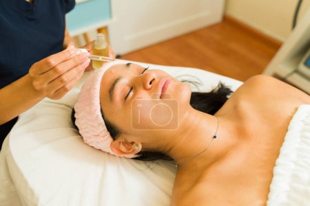 Foto de Mujer hispana joven relajándose mientras disfruta de un tratamiento facial usando aceite de ácido hialurónico en el spa de belleza - Imagen libre de derechos