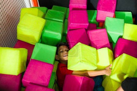 Foto de Chico latino sonriendo mientras se divierten mucho jugando con cubos de espuma de colores en el foso de la sala de juegos - Imagen libre de derechos
