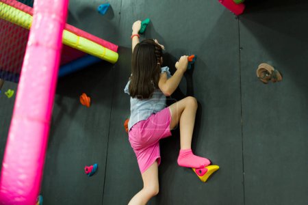 Foto de Niña activa que se divierte escalando en roca durante su tiempo de recreación y jugando juegos en la sala de juegos cubierta - Imagen libre de derechos