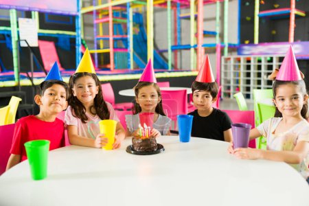 Photo for Cheerful group of children celebrating their friends birthday party with a cake wearing hats in a beautiful indoor playground - Royalty Free Image