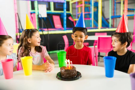 Foto de Niño hispano celebrando una fiesta de cumpleaños de niños con sus amigos y cantando feliz cumpleaños comiendo pastel en la sala de juegos - Imagen libre de derechos