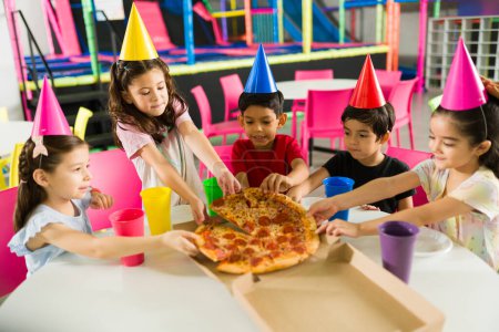 Foto de Feliz grupo de niños comiendo deliciosa pizza mientras celebran una fiesta de cumpleaños y se divierten juntos en la sala de juegos - Imagen libre de derechos