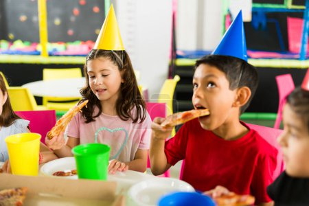 Photo for Fun group of children eating delicious pizza and cake while celebrating a friends birthday party together in the playground - Royalty Free Image