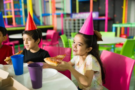 Foto de Hermosa niña sonriendo mientras come pizza y celebrando su fiesta de cumpleaños con amigos en el patio de recreo - Imagen libre de derechos