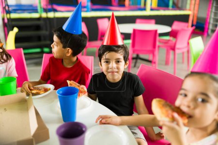 Foto de Hermoso grupo de niños riendo y disfrutando comiendo pizza durante una divertida fiesta de cumpleaños con amigos en el patio de recreo - Imagen libre de derechos
