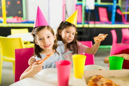 Foto de Adorables niñas y amigos abrazándose mientras usan sombreros de fiesta y comiendo pizza con pastel celebrando un cumpleaños de amigos - Imagen libre de derechos