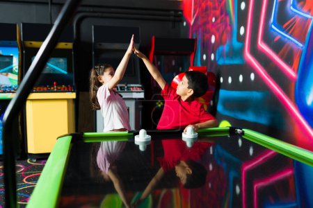 Photo for Excited kids doing a high five and celebrating winning a competition of air hockey with friends in the arcade playroom - Royalty Free Image