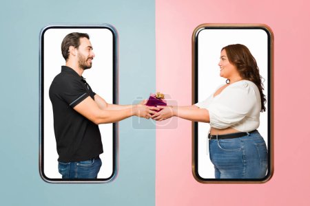 Photo for Beautiful boyfriend and girlfriend on a virtual date giving a present against a smartphone screen and having a long-distance relationship - Royalty Free Image