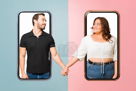 Photo for Beautiful happy couple holding hands meeting on an online dating app or having a long-distance relationship against a smartphone screen - Royalty Free Image