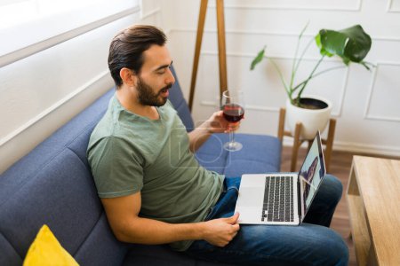 Téléchargez les photos : Beau homme se détendre à la maison boire du vin et avoir une date virtuelle et profiter d'un appel vidéo en ligne sur l'ordinateur portable - en image libre de droit