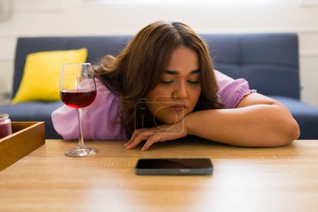 Foto de Mujer latina que parece triste y deprimida con una copa de vino esperando la llamada de su novio por teléfono mientras lo echa de menos - Imagen libre de derechos