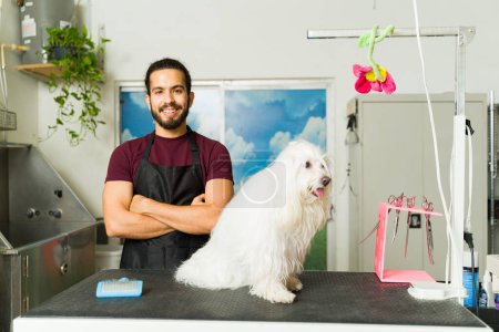 Téléchargez les photos : Heureux homme hispanique souriant tout en travaillant comme toiletteur au spa pour chiens et en donnant un bain et une coupe de cheveux à un chien maltais - en image libre de droit