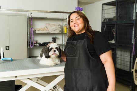 Téléchargez les photos : Portrait de femme latine travaillant comme toiletteur animal souriant posant avec un chien shih tzu adorable après un bain et une coupe de cheveux - en image libre de droit