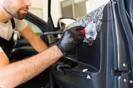 Foto de Primer plano de un joven limpiando el panel interior de la puerta del coche con jabón con cepillo en su trabajo en el servicio de detalle automático - Imagen libre de derechos