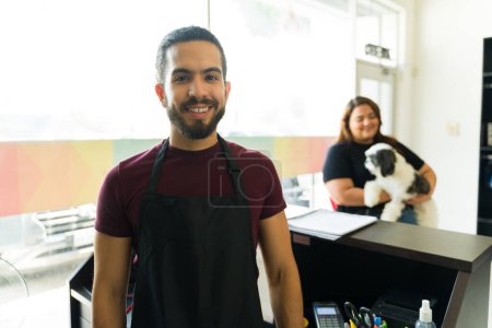 Foto de Hombre latino guapo y peluquero de mascotas sonriendo mientras disfruta trabajando en la tienda de spa de aseo cuidando perros hermosos - Imagen libre de derechos