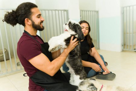 Foto de Joven latino abrazando y acariciando a un hermoso perro shih tzu mientras trabaja como peluquero de mascotas u hotel de guardería - Imagen libre de derechos