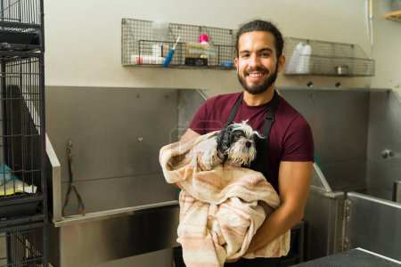 Téléchargez les photos : Attrayant toiletteur heureux animal souriant tout en portant un chien shih tzu dans une serviette après un bain pour se brosser les cheveux - en image libre de droit