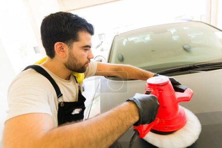 Photo for Attractive young male employee using orbital polisher for waxing a car body at auto detail service - Royalty Free Image